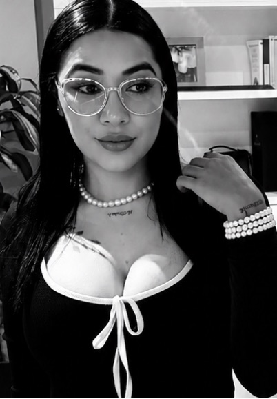 A woman with long dark hair and glasses wearing a pearl necklace, looking into the camera in a black-and-white portrait